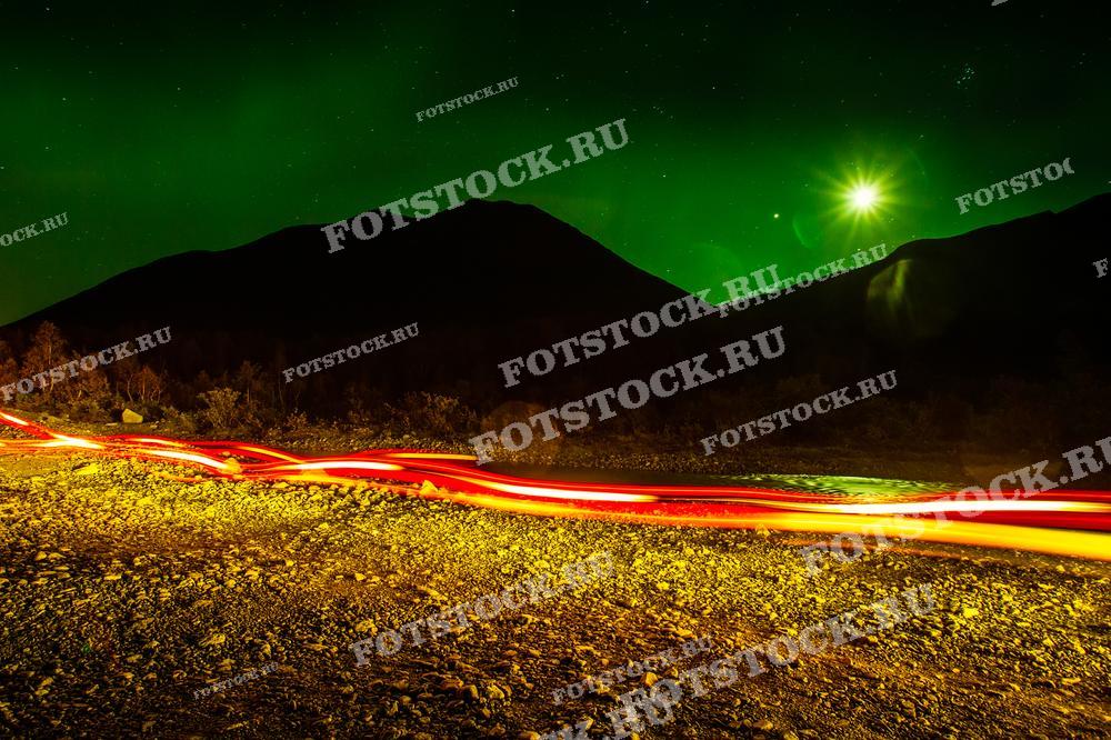 На фотографии изображено северное сияние над горами Хибин в Мурманской области. Яркие зеленые лучи сияния заполняют небо, а на переднем плане длинные световые следы от движущихся автомобилей добавляют динамику к статичному природному пейзажу. Луна, освещающая горизонт, и сияние создают атмосферу мистической северной ночи. Это уникальное сочетание природных и человеческих элементов подчёркивает красоту северного региона.